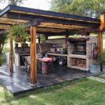 Covered outdoor cooking area with brick grill, wooden beams, and dining table on grass.
