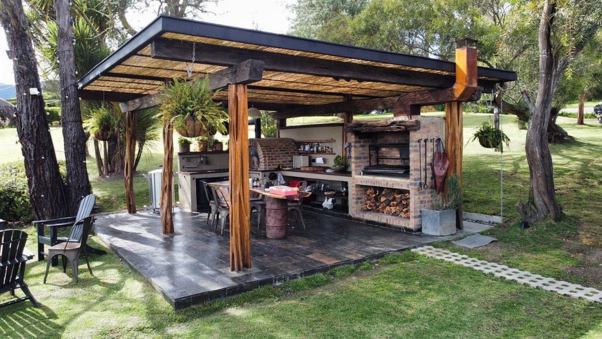 Covered outdoor cooking area with brick grill, wooden beams, and dining table on grass.