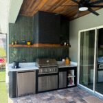 Outdoor kitchen with dark green tiled backsplash, stainless steel grill, and wooden ceiling.