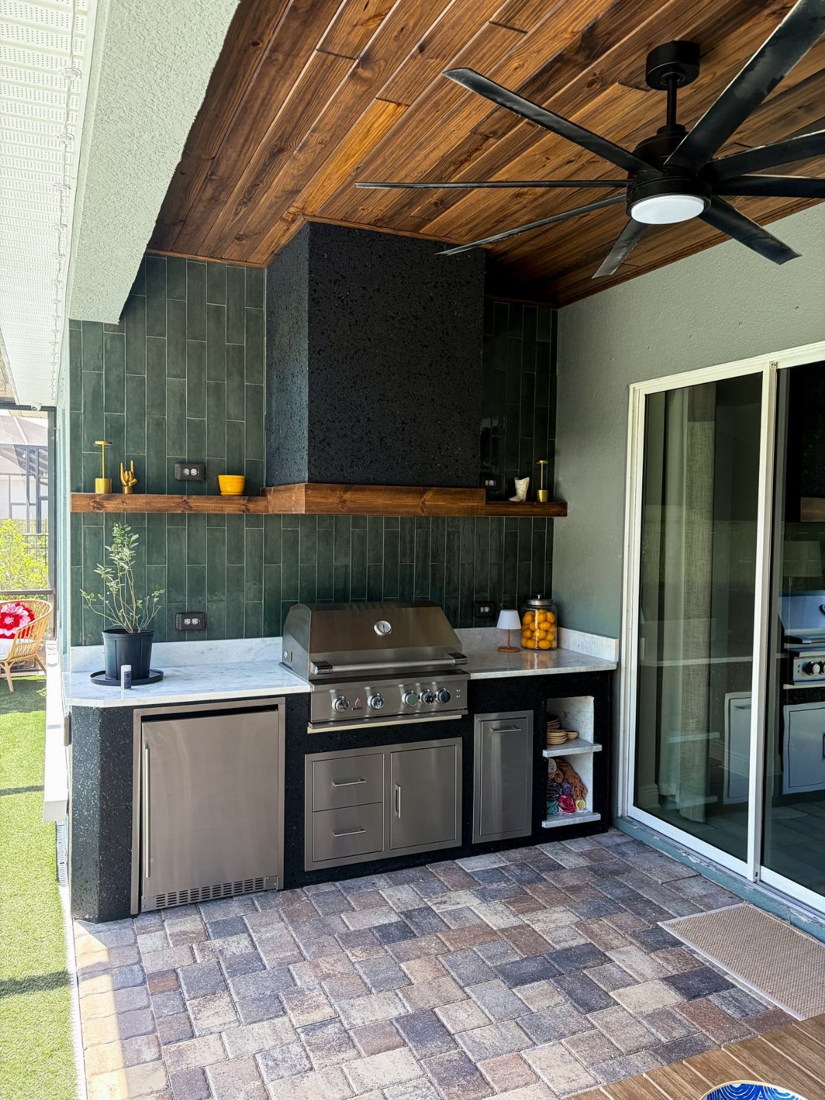 Outdoor kitchen with dark green tiled backsplash, stainless steel grill, and wooden ceiling.