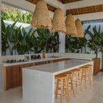 Outdoor kitchen with white concrete island, wooden stools, and straw pendant lights.