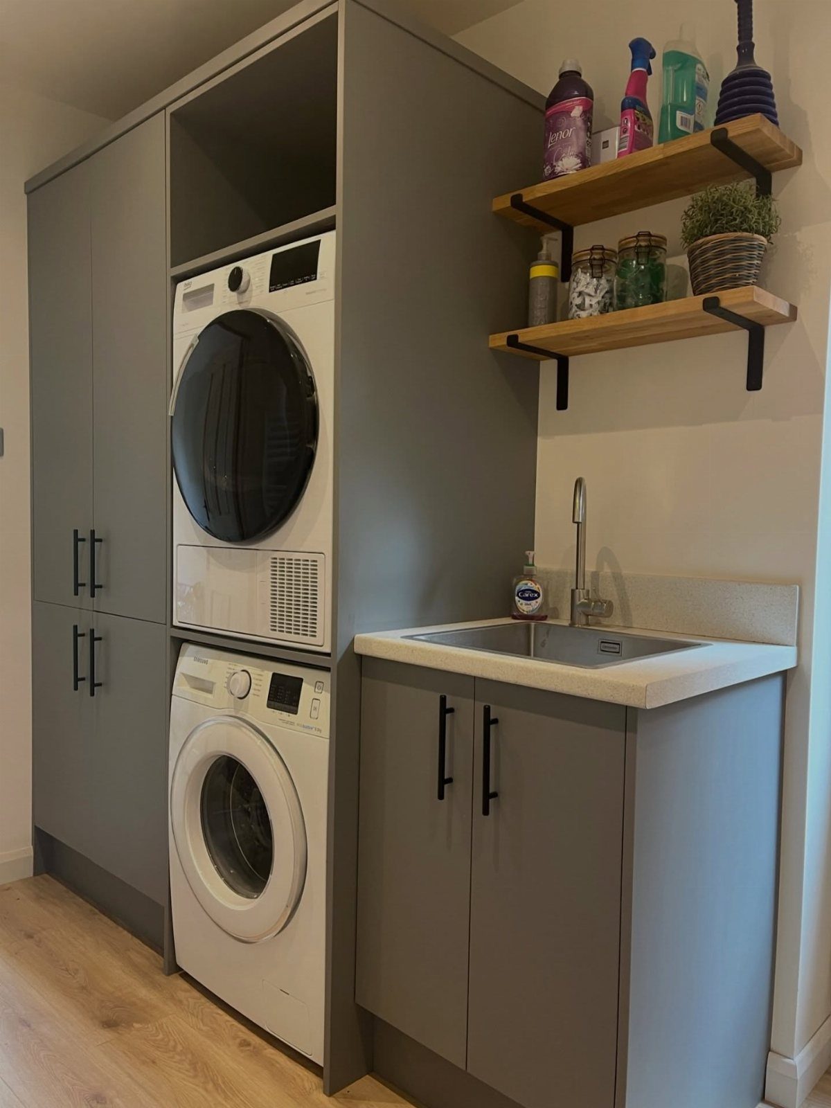 Compact utility kitchen area with grey cabinets, stacked laundry machines, and floating shelves.